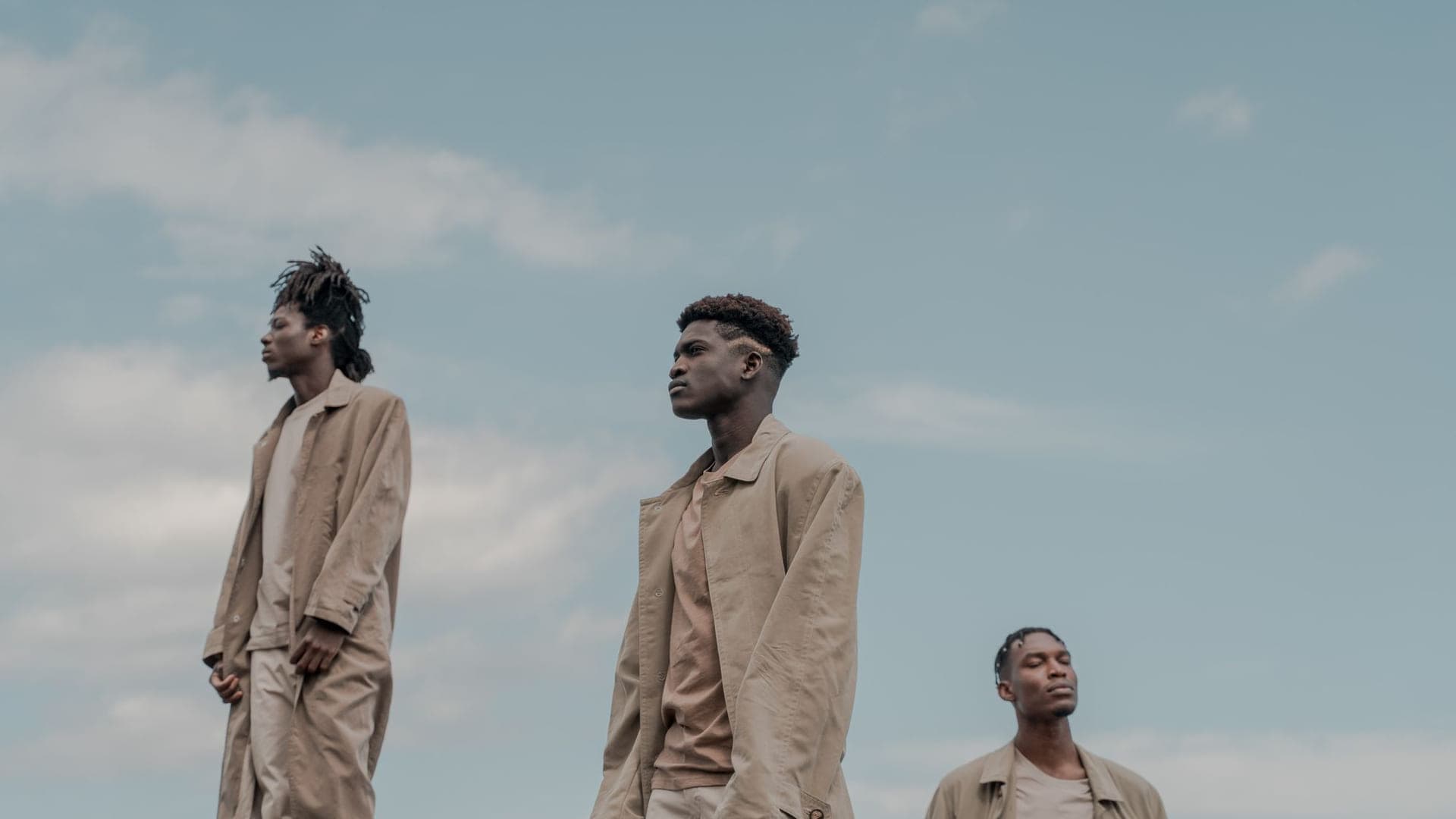 Three men in long coats against an open sky