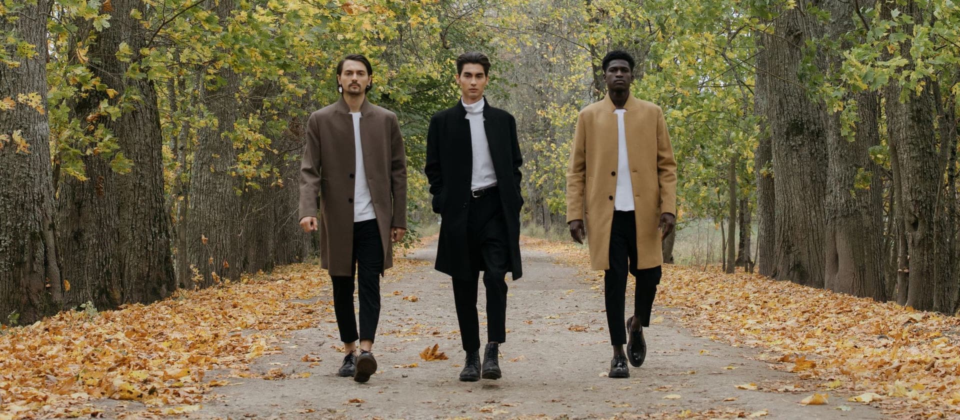 Three men in layered autumn coats walking a leaf-lined path